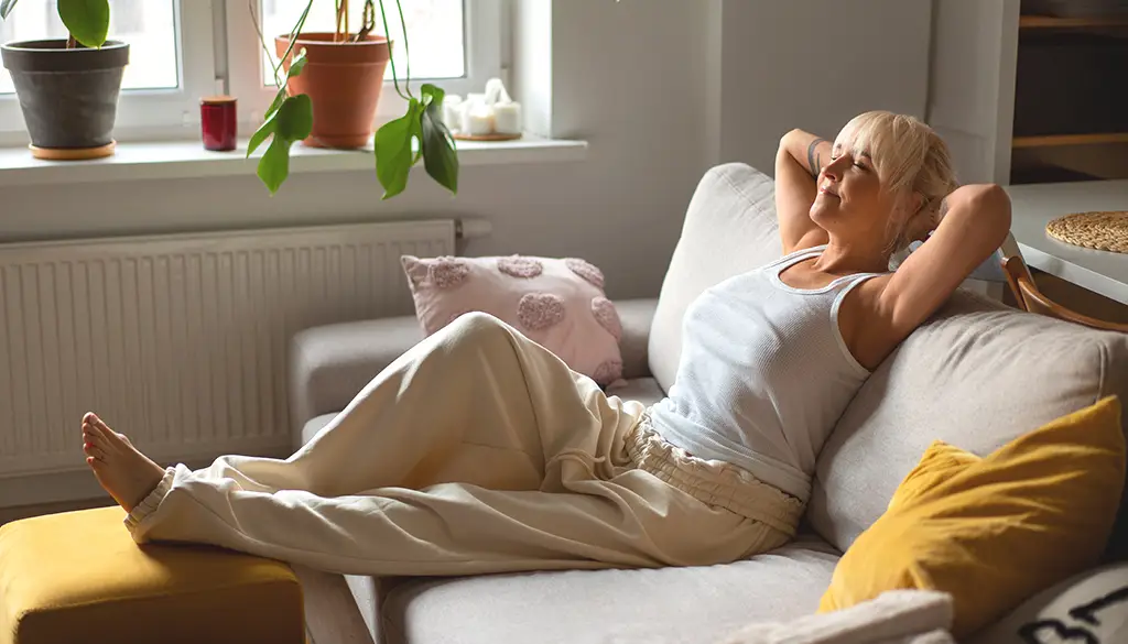 Woman meditating on a couch at home with eyes closed, arms behind her head, in a comfortable everyday setting.