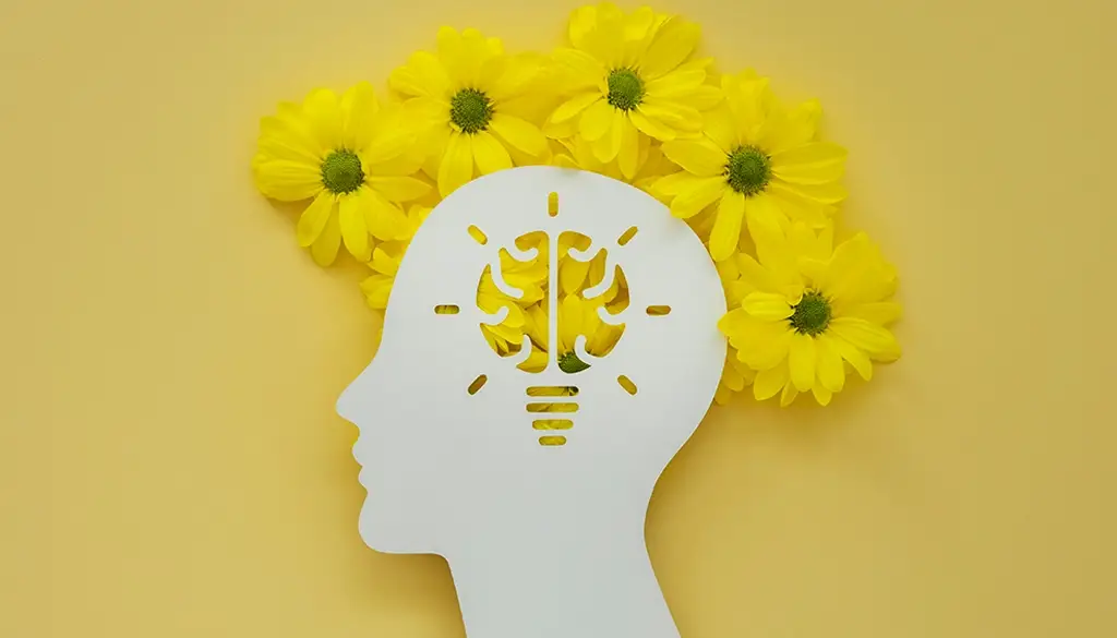 White paper cutout of a human head with a brain-shaped lightbulb at its center, surrounded by yellow flowers — symbolizing insight, clarity, and the bloom of a new idea.